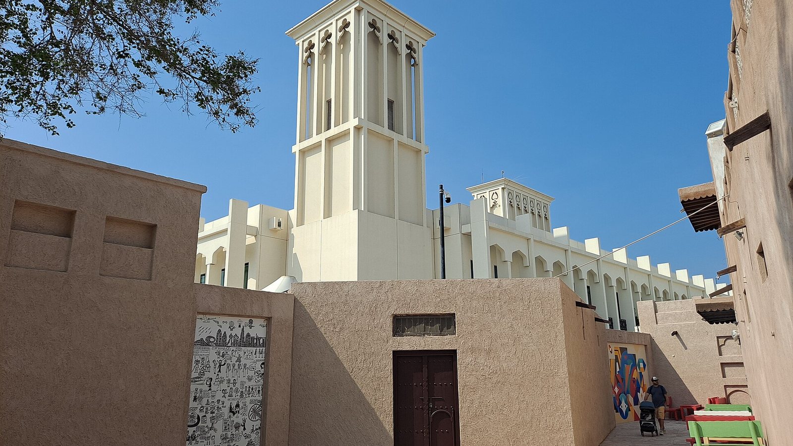 A cream-coloured wind tower rising above sand-coloured walls in Dubai's Al Fahidi Historical Neighbourhood, under a clear blue sky