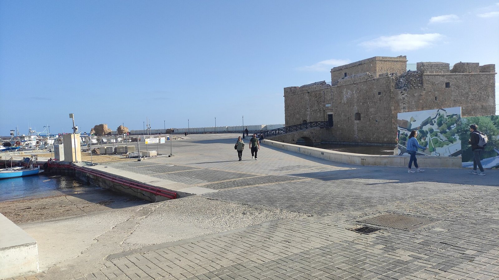 The stone Paphos Castle at the end of the harbour promenade in Paphos, with people walking under a clear blue sky