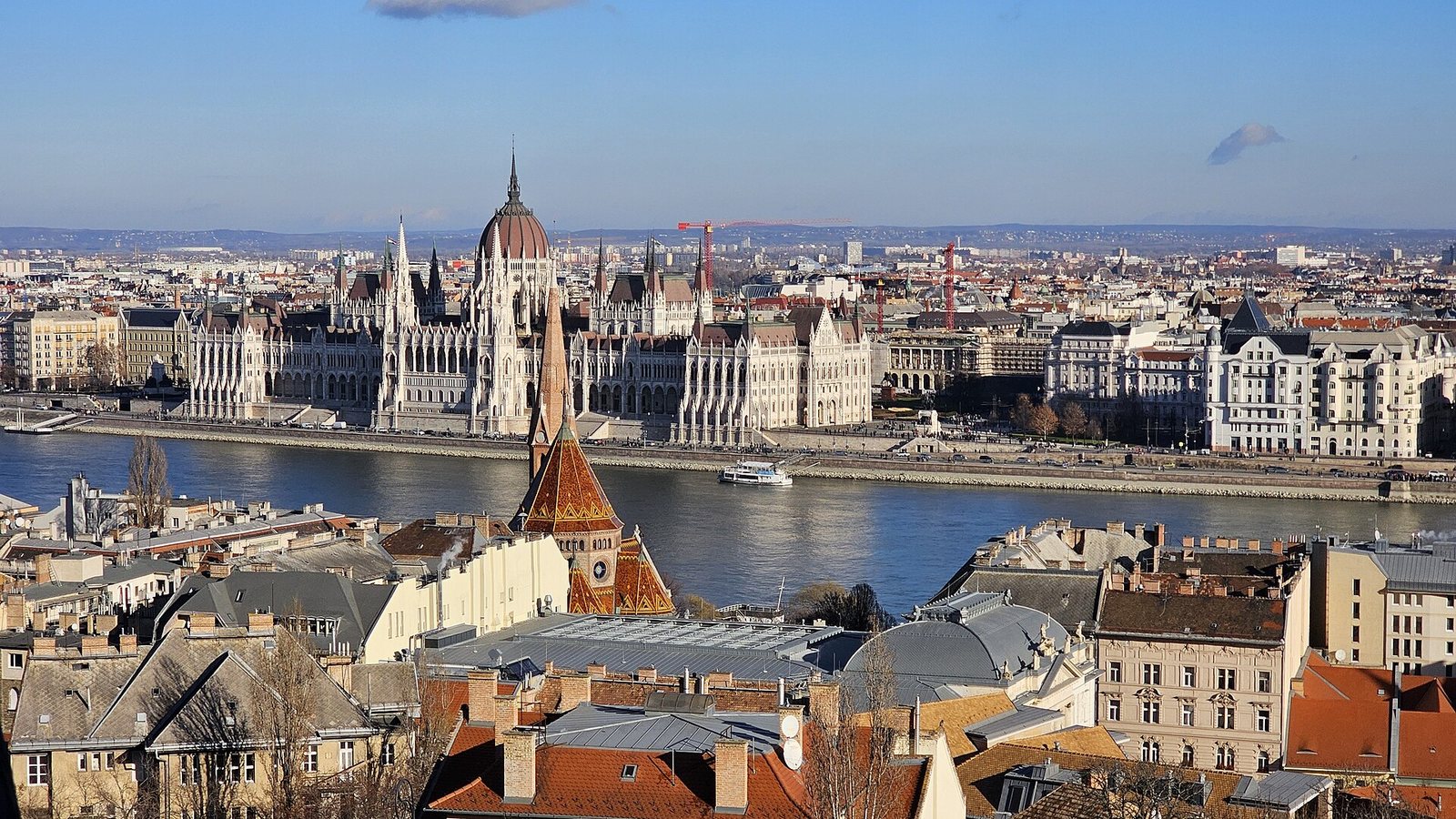 The Hungarian Parliament Building on the bank of the Danube in Budapest, seen across the river above red-roofed buildings under a clear sky