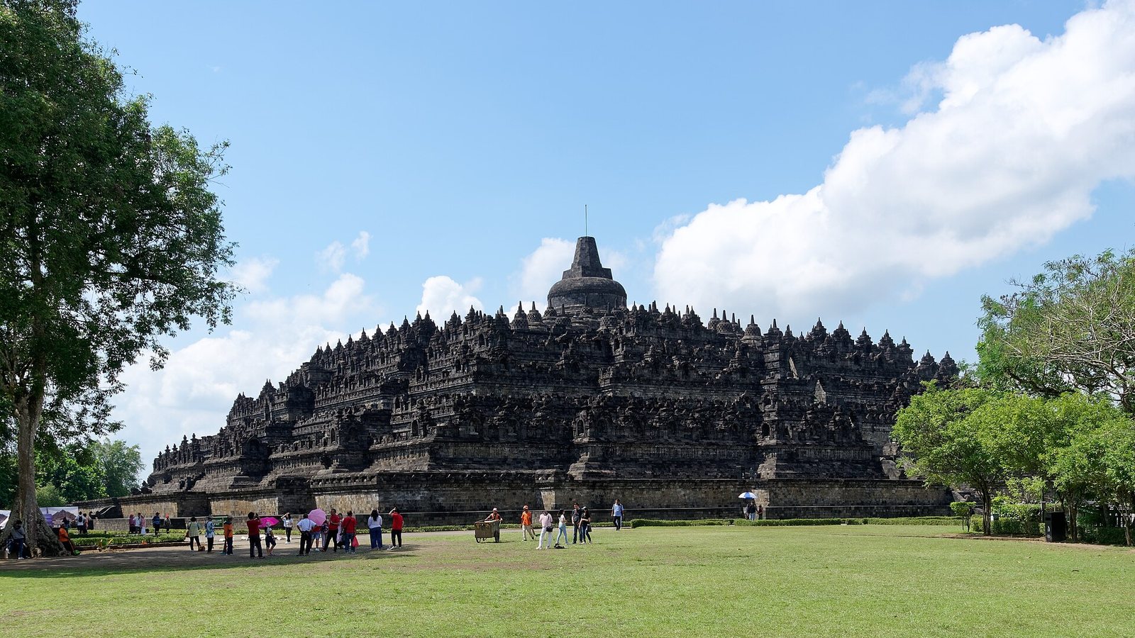 The stone stupas and terraces of Borobudur temple in Central Java under a blue sky, with visitors on the lawn in front