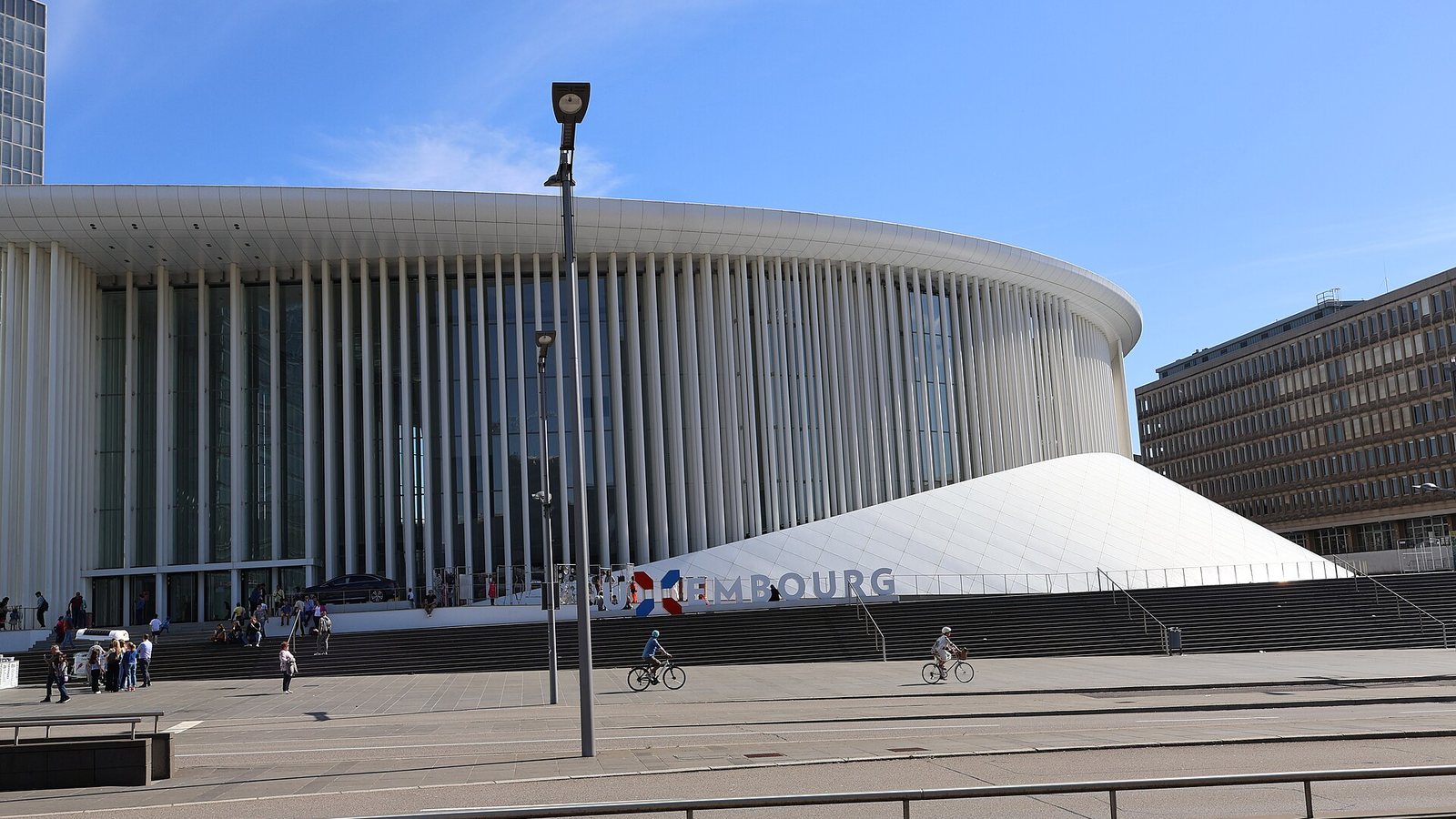 The white colonnaded Philharmonie concert hall on the Kirchberg plateau in Luxembourg City, with cyclists crossing the square in front under a blue sky