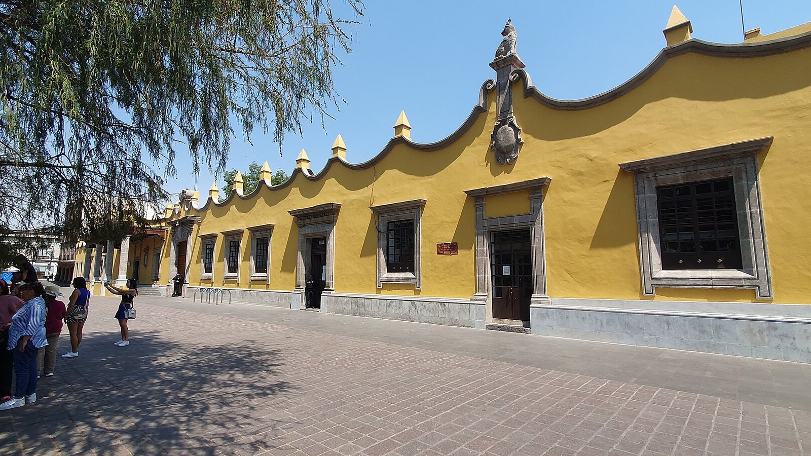 Long yellow colonial building with stone window frames and decorative crenellations along a paved plaza under a clear blue sky, with a tree and pedestrians on the left
