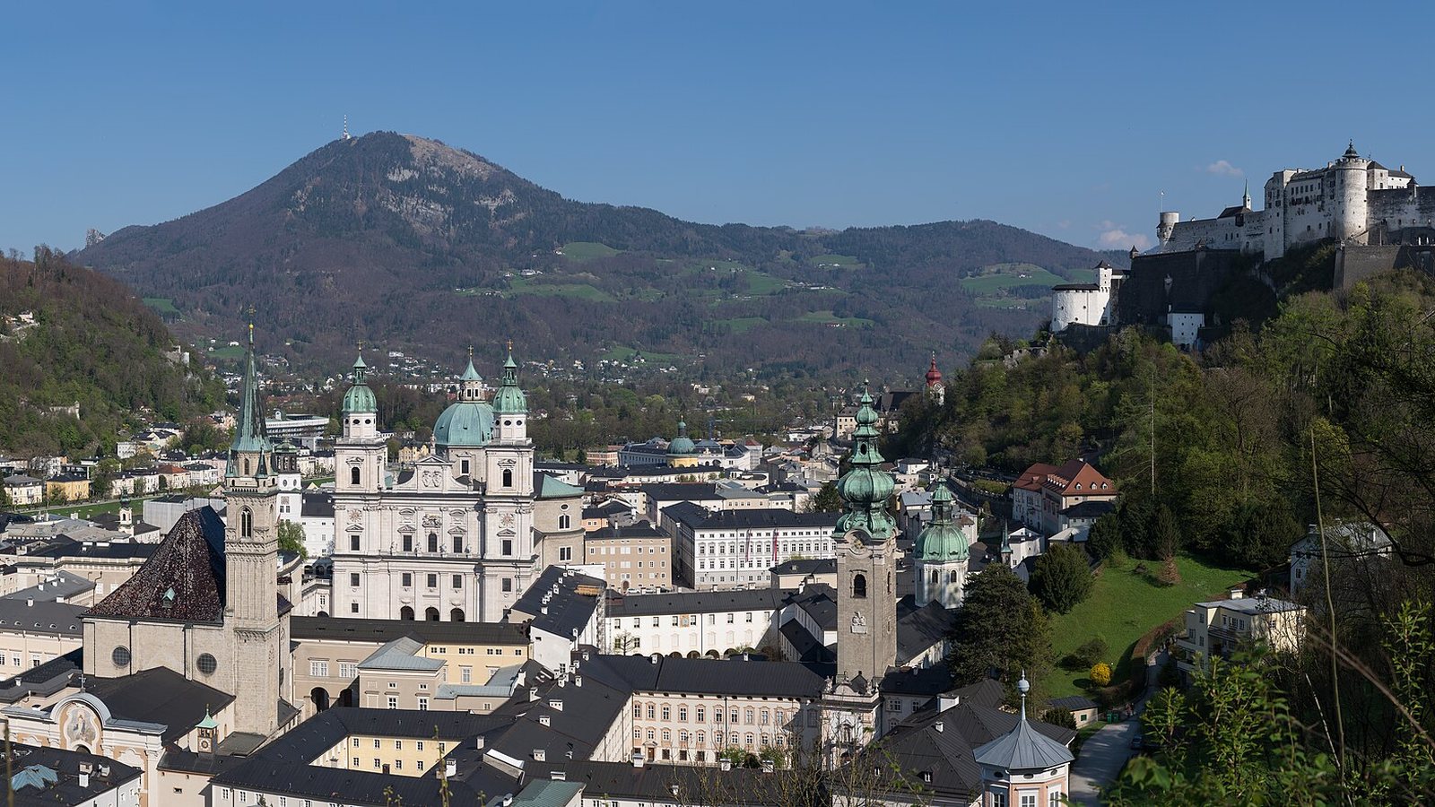 Salzburg old town with the cathedral domes and green church spires below Hohensalzburg Fortress, with the Gaisberg mountain behind under a blue sky