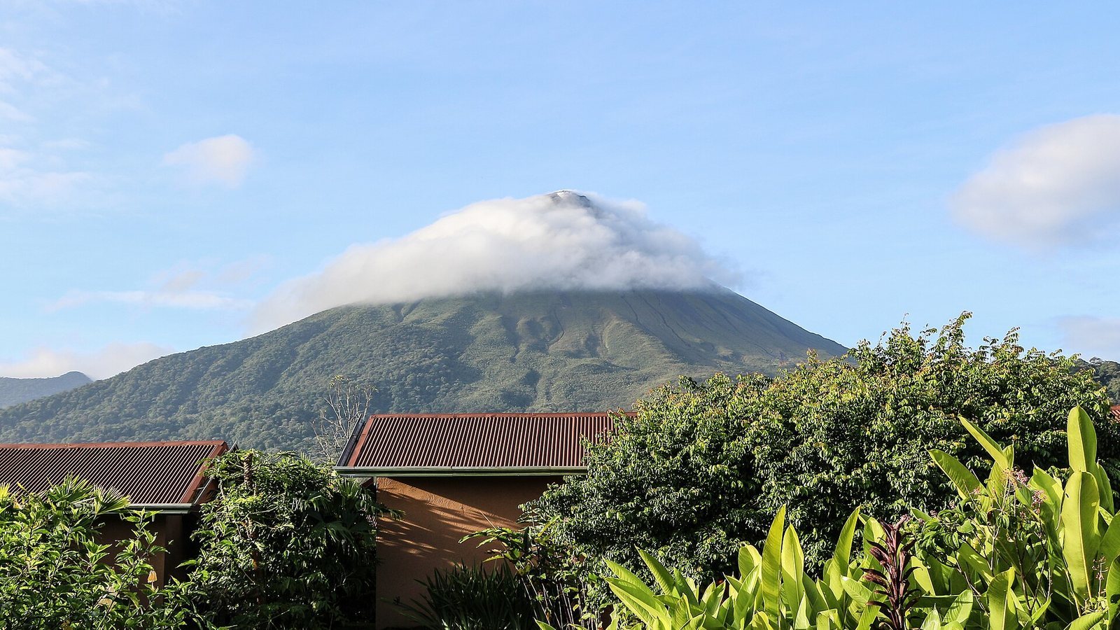 Arenal Volcano in Costa Rica with a cloud cap over its peak above green forest and rooftops
