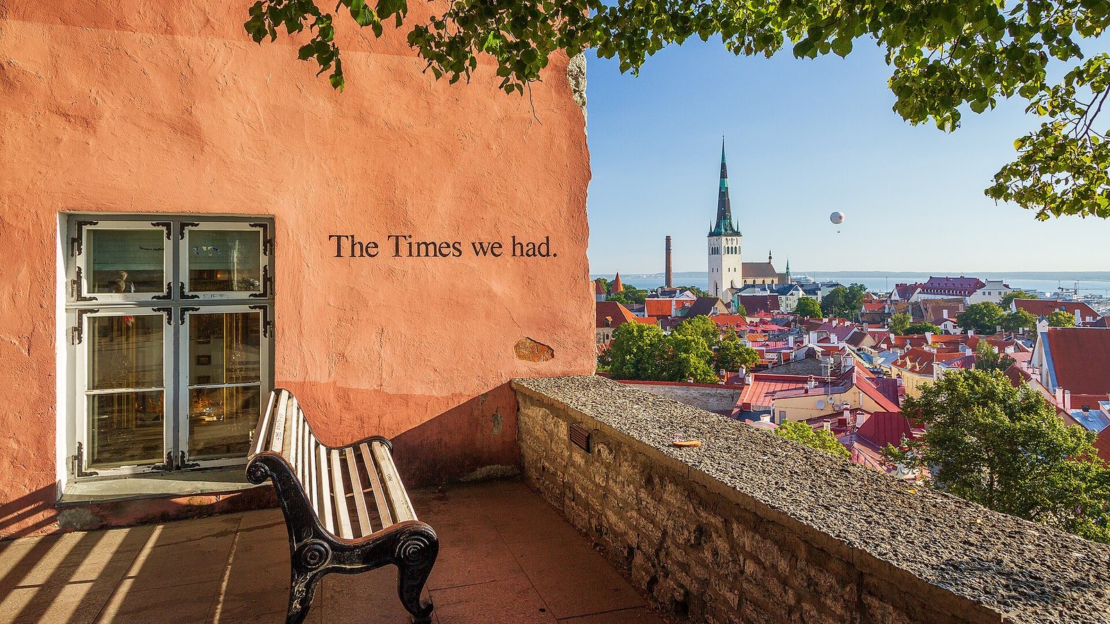 The Kohtuotsa viewing platform in Tallinn with a bench beside an orange wall, looking over red roofs of the Old Town to St Olaf's Church and the sea