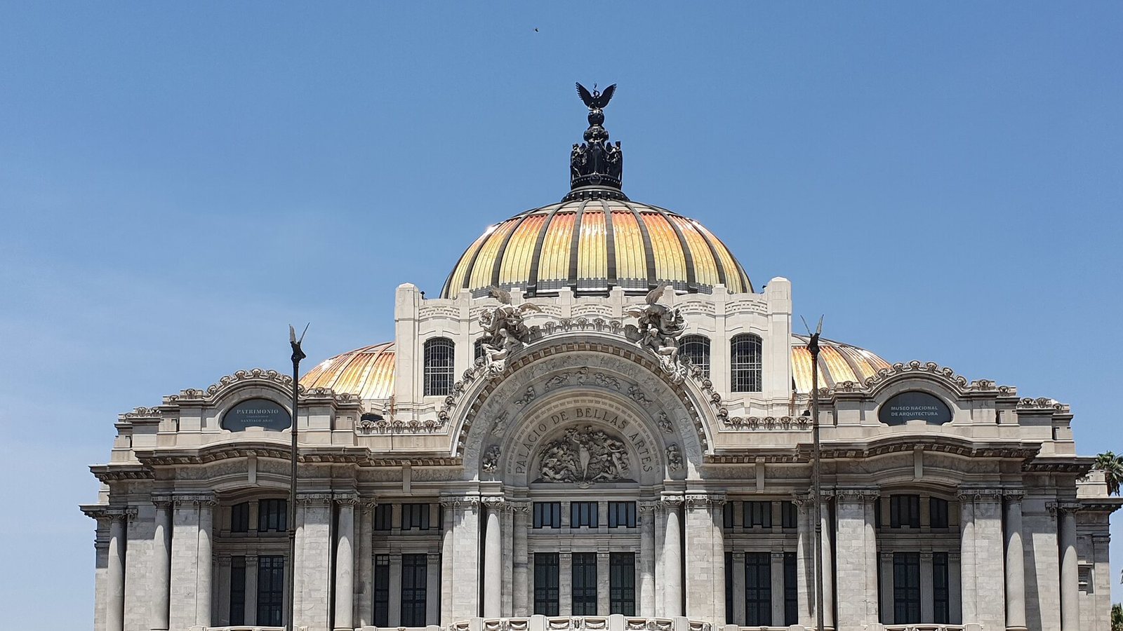 White marble facade of the Palacio de Bellas Artes in Mexico City with its orange and yellow tiled dome and a bronze eagle sculpture on top against a clear blue sky