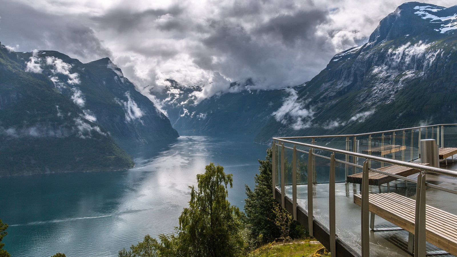 View from a railed viewing platform down the Geirangerfjord between steep green mountains with low cloud and snow patches
