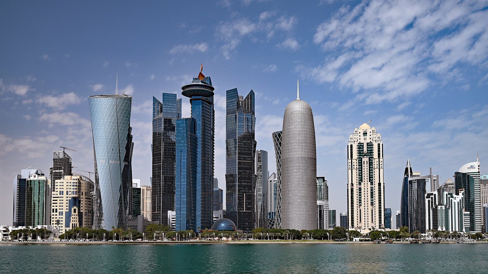 Glass towers of the West Bay district in Doha rising behind the Corniche waterfront under a blue sky with light cloud