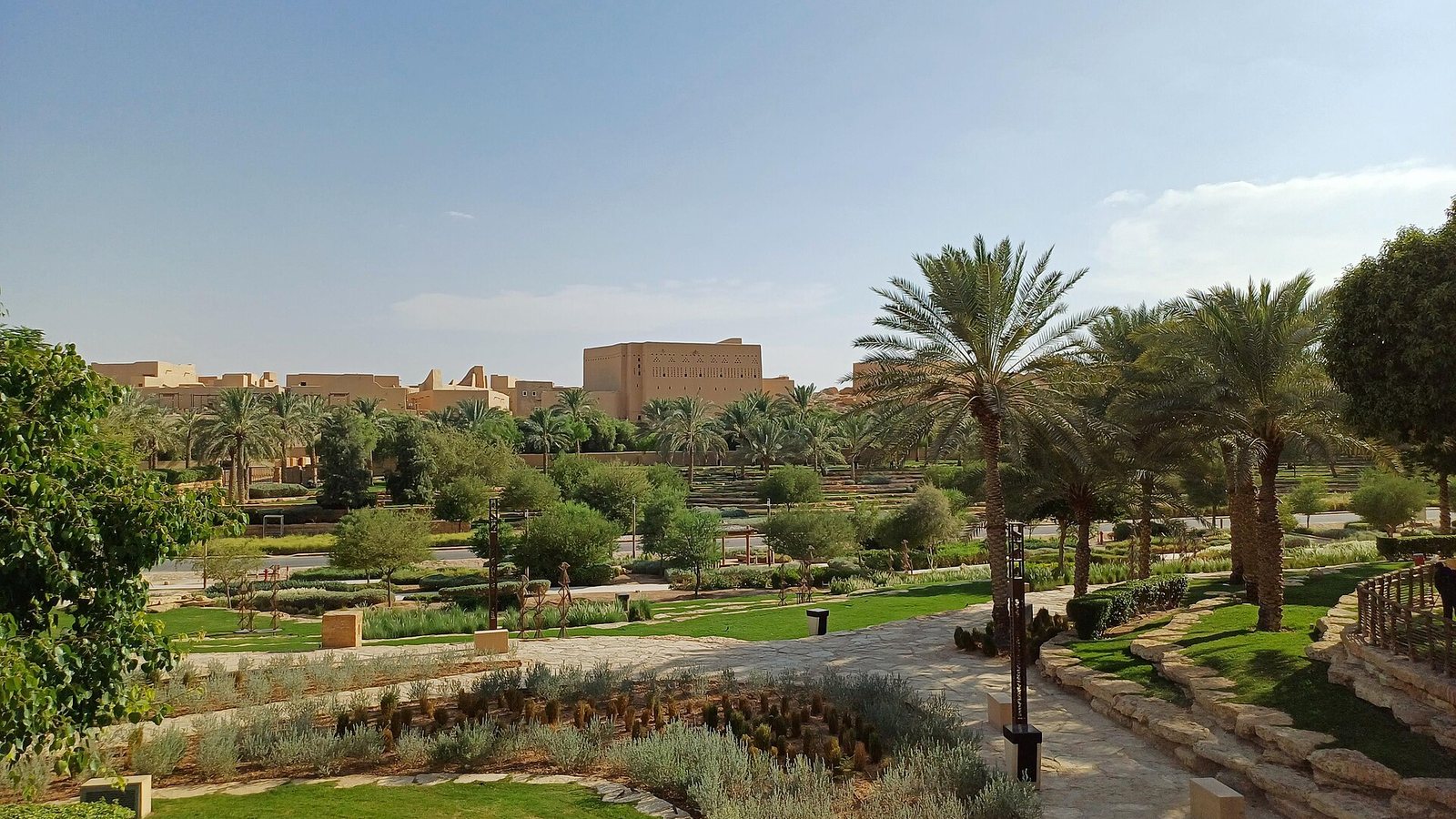 Landscaped gardens with palm trees, lawns and stone paths in Diriyah, with mud-brick style buildings of At-Turaif on the skyline under a pale blue sky