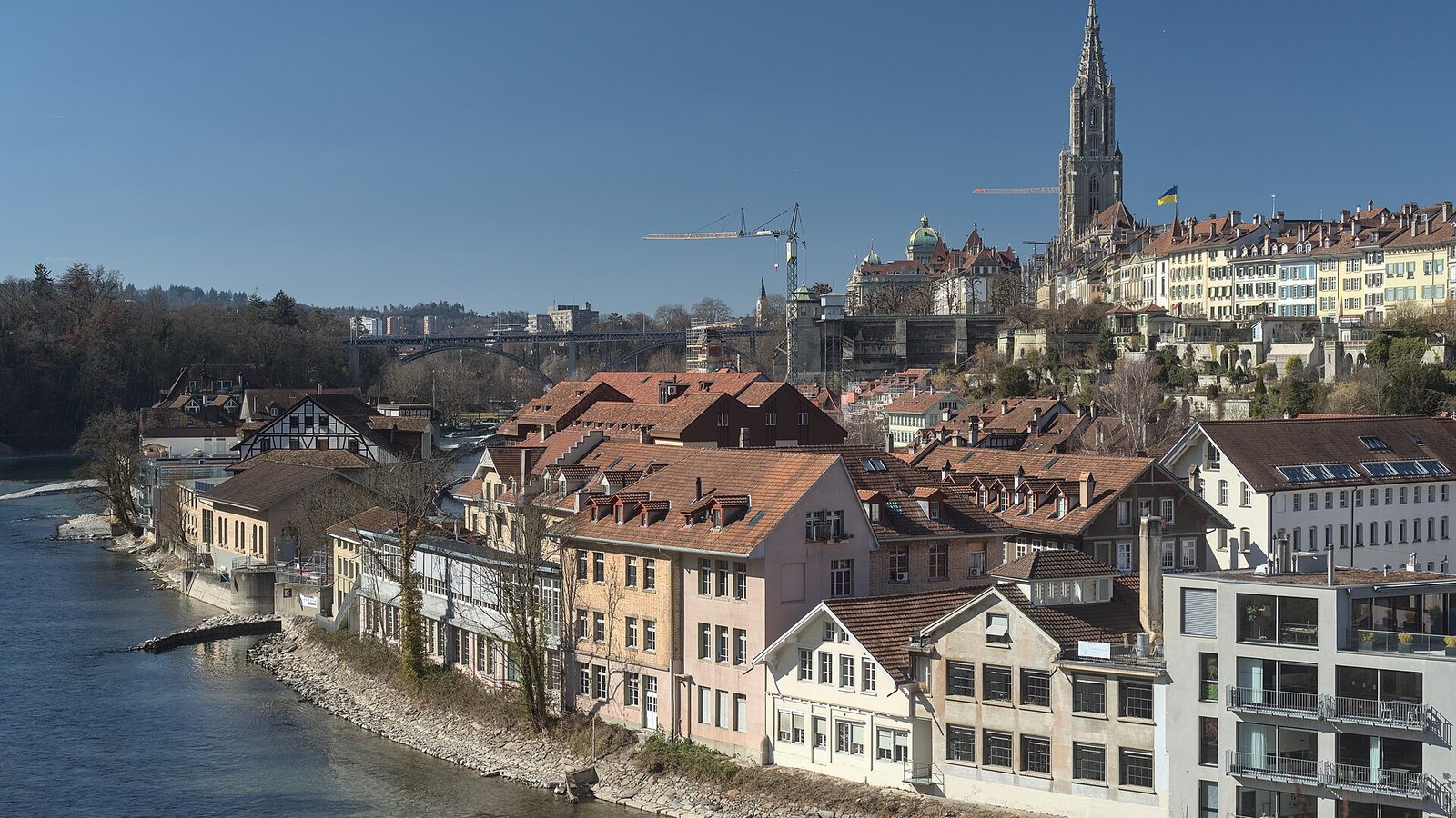 The old town of Bern rising above the river Aare, with the spire of Bern Minster and rows of townhouses under a clear blue sky