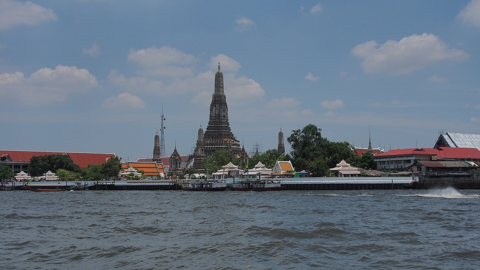 Wat Arun, the Temple of Dawn, seen across the Chao Phraya River in Bangkok under a blue sky with scattered clouds