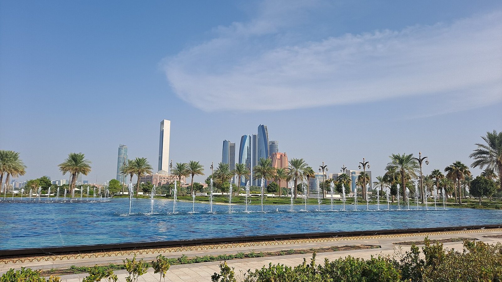 Fountains in the gardens of Qasr Al Watan with the towers of the Abu Dhabi skyline behind, under a blue sky