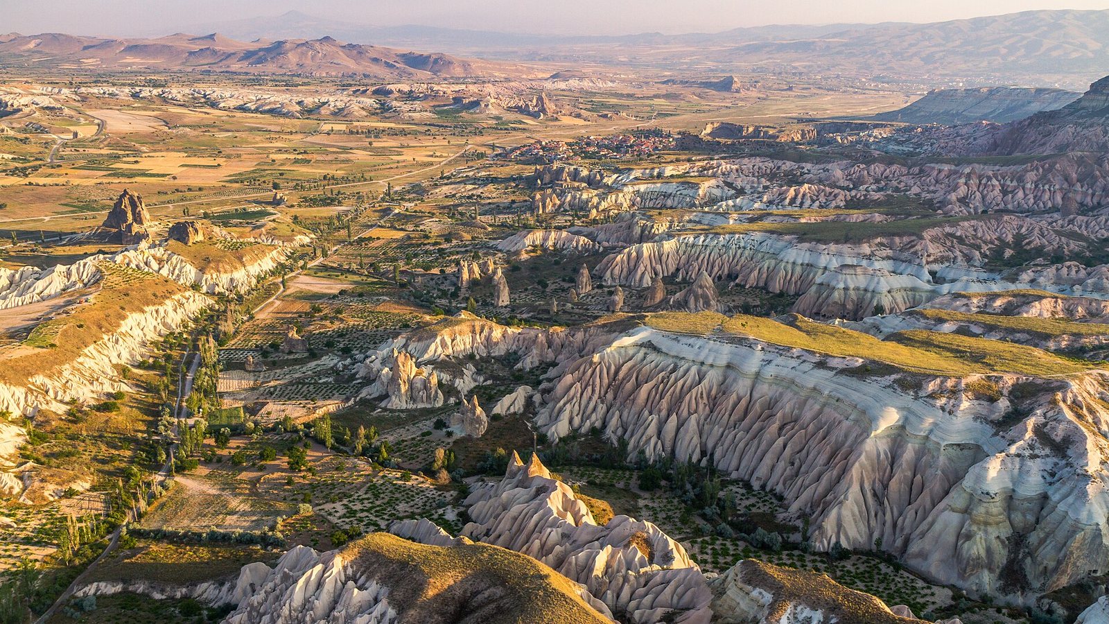 Aerial view over Cappadocia with eroded white and pink rock valleys, fields, a village and distant mountains in warm light