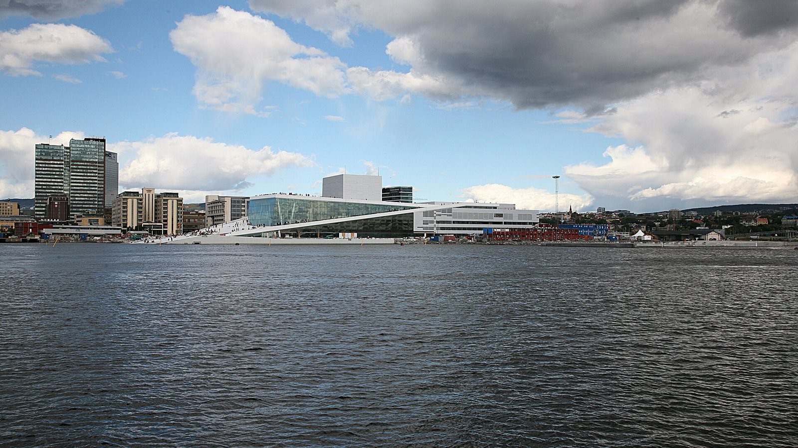 The white sloping Oslo Opera House at Bjørvika on the harbour, with office towers behind and a sky of white and grey clouds