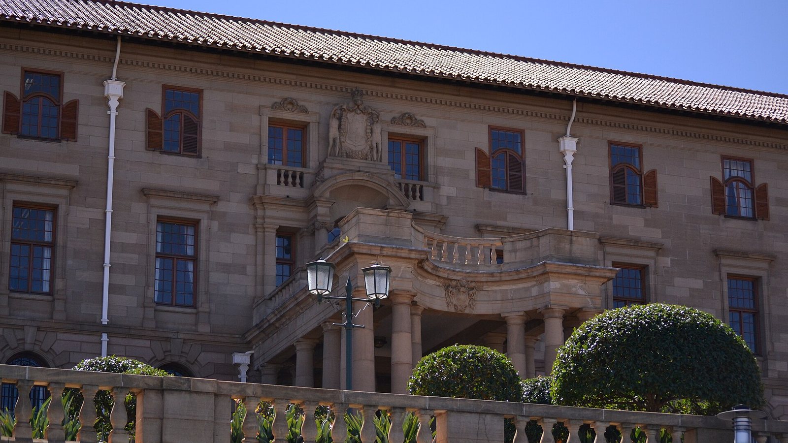 Sandstone facade of the Union Buildings in Pretoria with tiled roof, shuttered windows, a carved coat of arms, a balustrade and clipped round shrubs under a blue sky