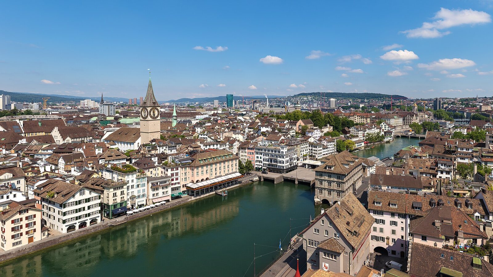 The old town of Zurich on both banks of the Limmat river with the clock tower of St. Peter church, seen from above under a blue sky