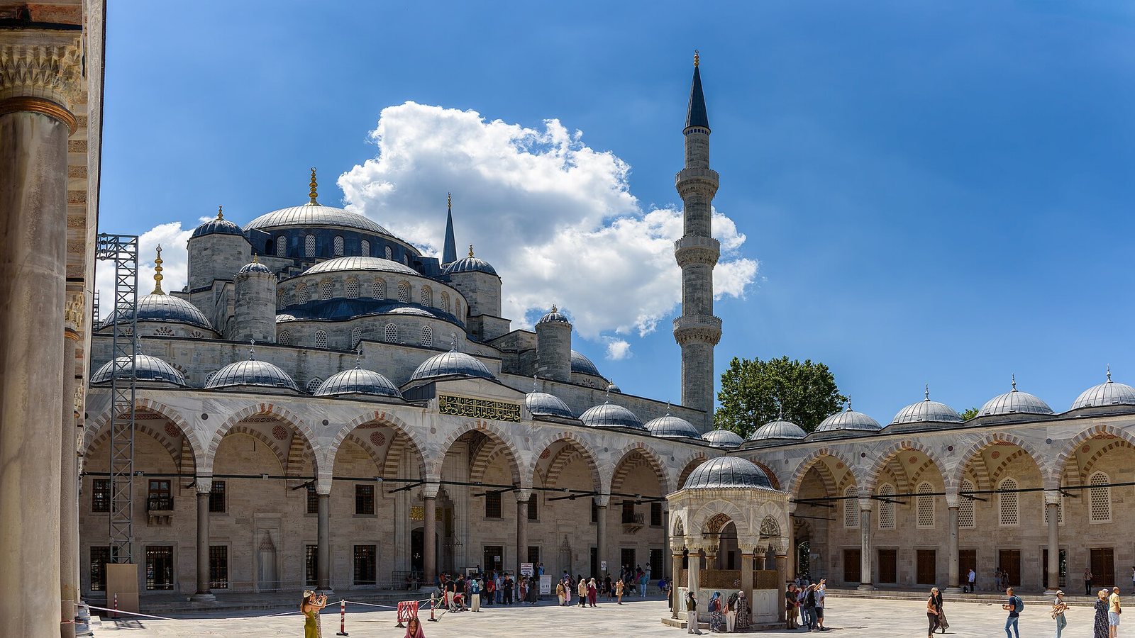 The domes and minaret of the Sultan Ahmed Mosque in Istanbul seen from its arcaded courtyard under a blue sky with a white cloud