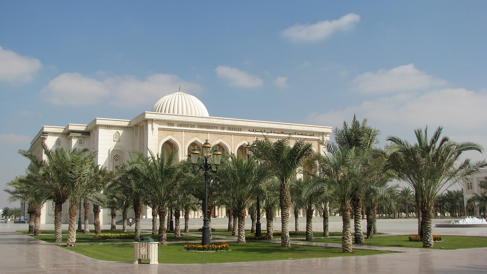The domed main building of the American University of Sharjah behind rows of date palms, under a blue sky