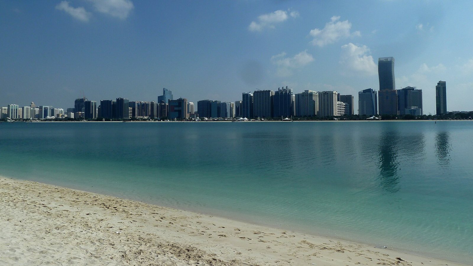 The Abu Dhabi skyline seen across turquoise water from a sandy beach on the Corniche