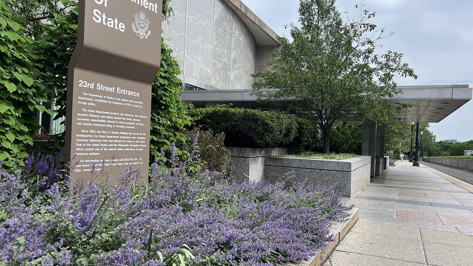 The 23rd Street entrance of the US Department of State building in Washington, with a sign and flowering plants